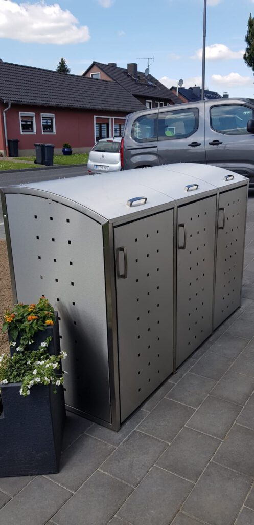 AT Garten- & Tiefbau Dreiteiliger Metallabfallbehälter mit Belüftungslöchern auf einem Gehweg.