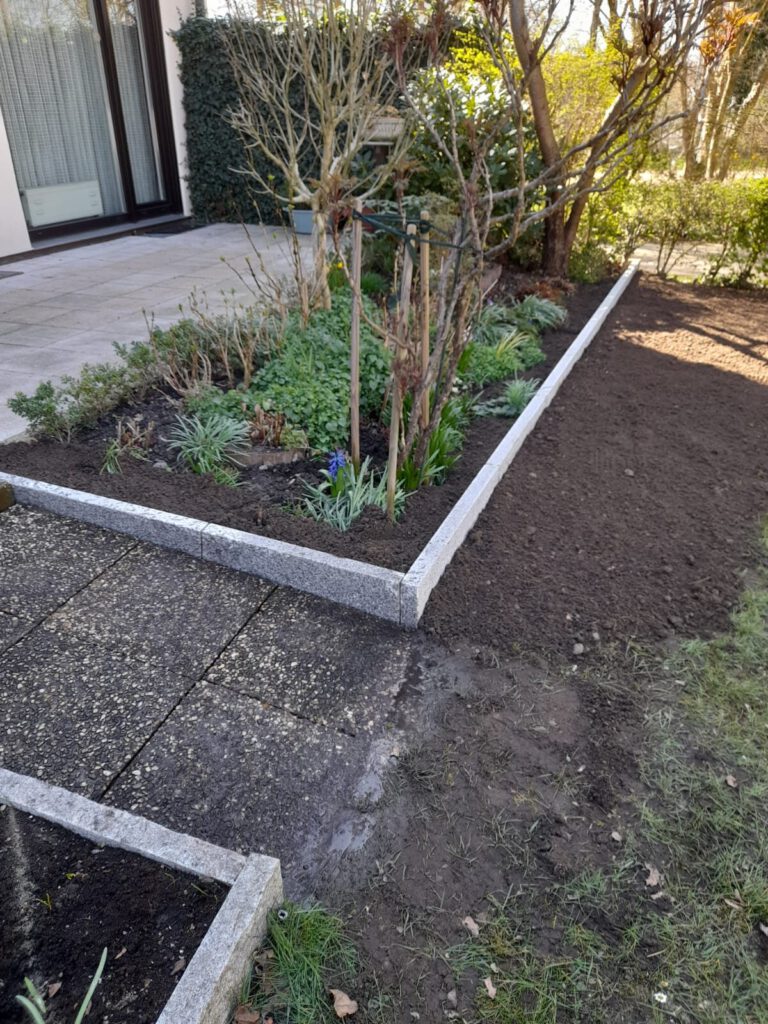 AT Garten- & Tiefbau Gartenbereich mit bepflanztem Beet, Steinrand und frisch bearbeitetem Boden.