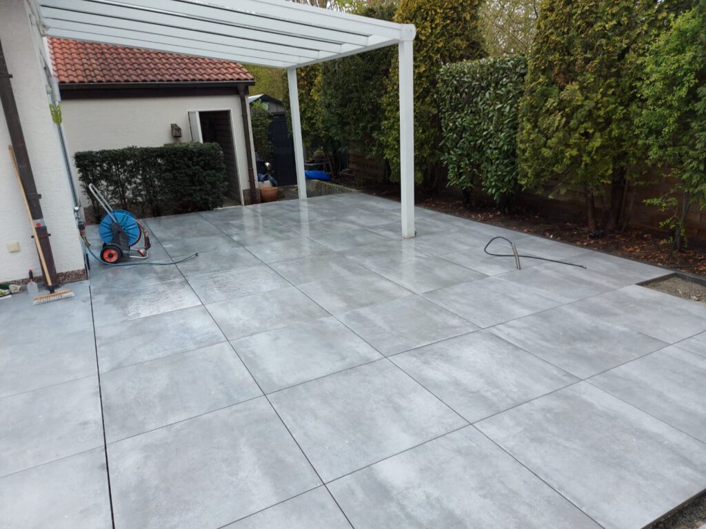 AT Garten- & Tiefbau Grau gefliester Außenbereich mit einer Überdachung und einem Gartenschlauch auf dem Boden.