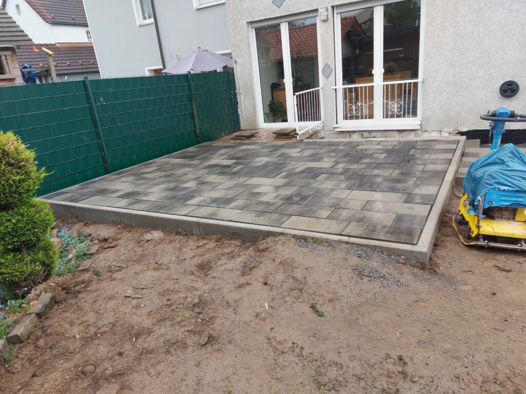 AT Garten- & Tiefbau Pflastersteinterrasse mit grauen Steinen hinter einem Wohnhaus, teilweise unvollständig.