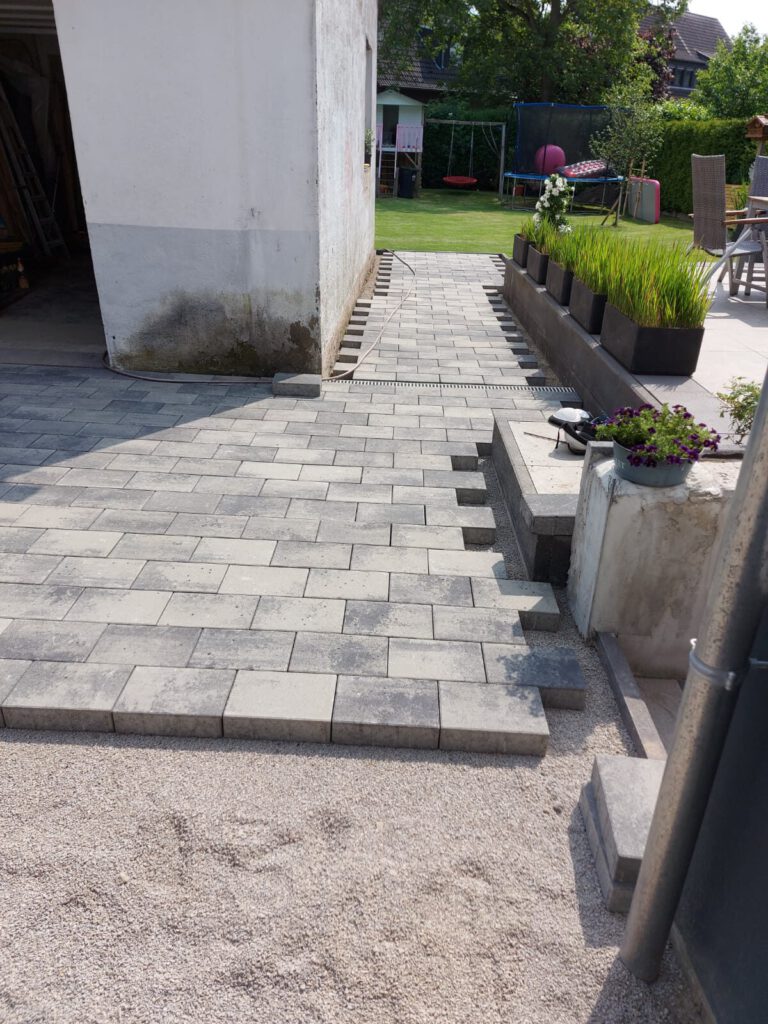 AT Garten- & Tiefbau Gepflasterter Weg aus grauen Betonsteinen, der zu einem Garten führt.