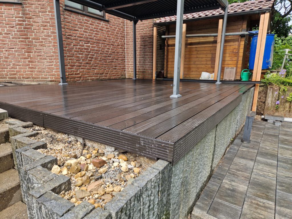 AT Garten- & Tiefbau Holzterrasse mit braunen Dielen und einer Steinmauer im Hintergrund.