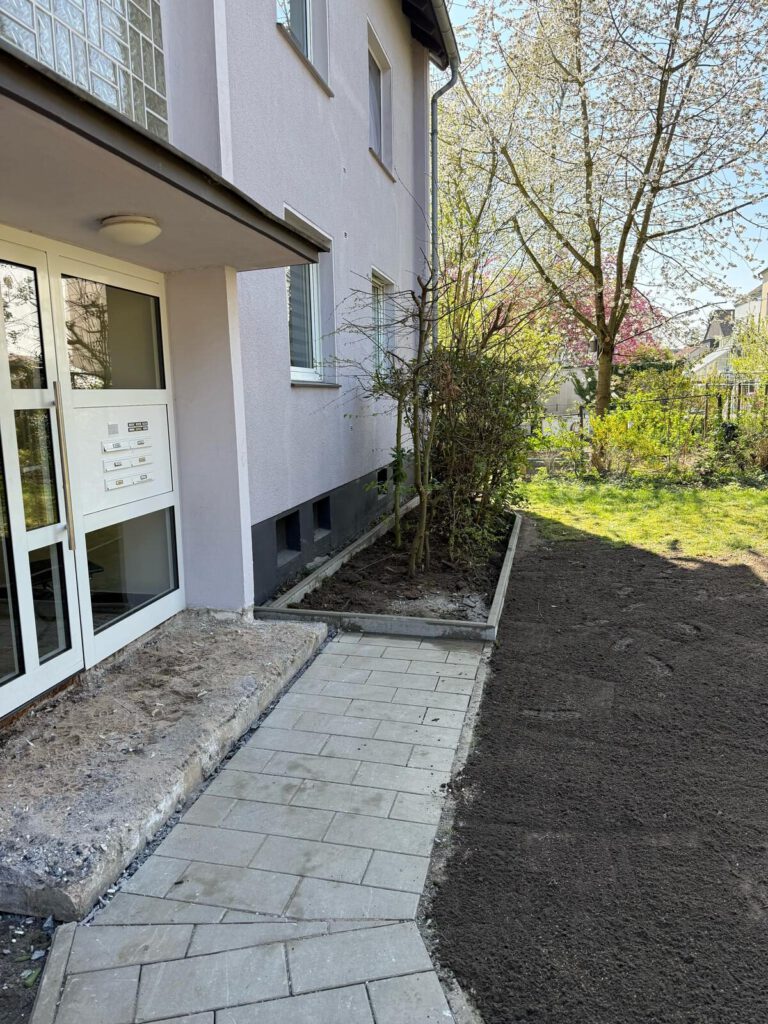 AT Garten- & Tiefbau Eingangsbereich eines Gebäudes mit neu verlegten Pflastersteinen und einer Bepflanzung.