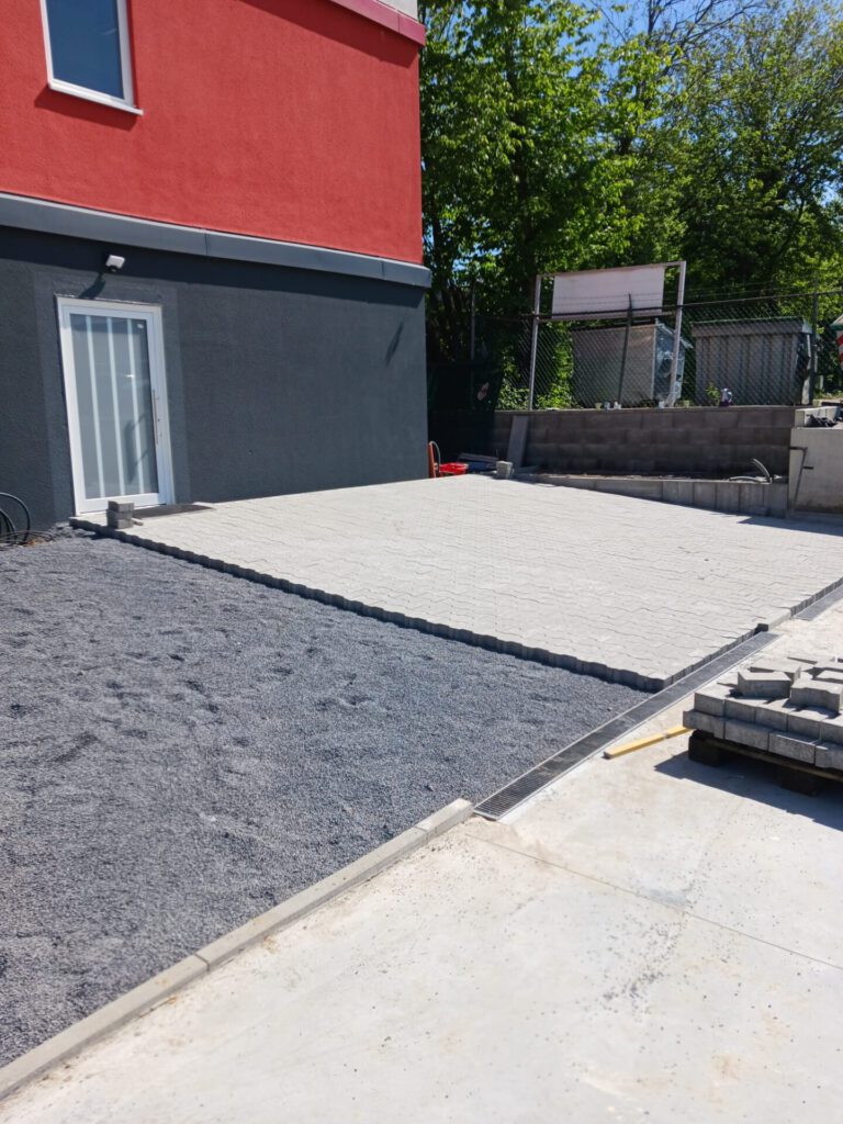 AT Garten- & Tiefbau Gepflasterte Fläche mit grauem Kies und hellen Betonplatten in einem Außenbereich.