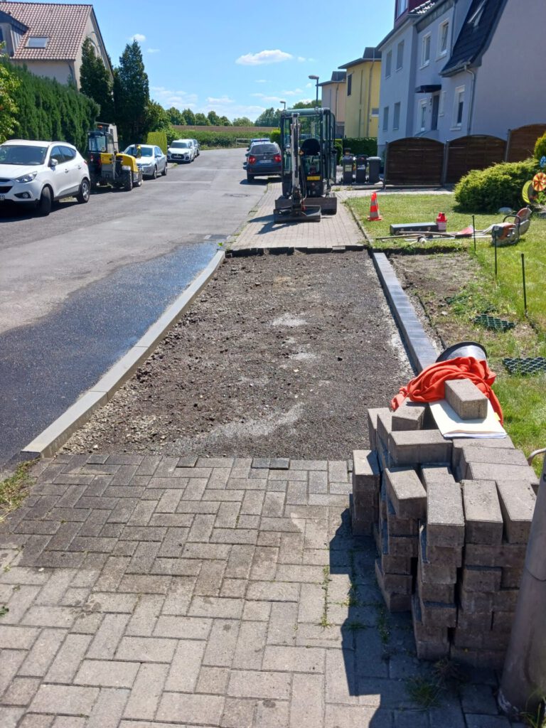 AT Garten- & Tiefbau Unvollendeter Gehweg mit Erde und Pflastersteinen auf einer Baustelle in einer Wohngegend.