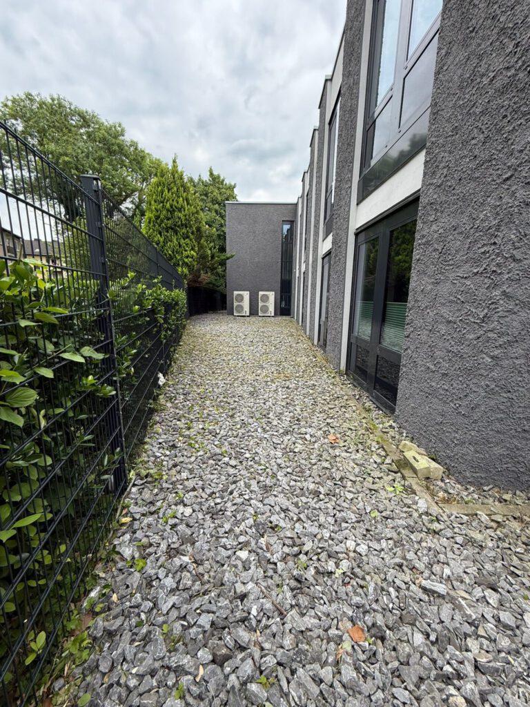AT Garten- & Tiefbau Ein Kiesweg zwischen einem Zaun und einer Gebäudefassade mit Fenstern und Klimaanlagen.