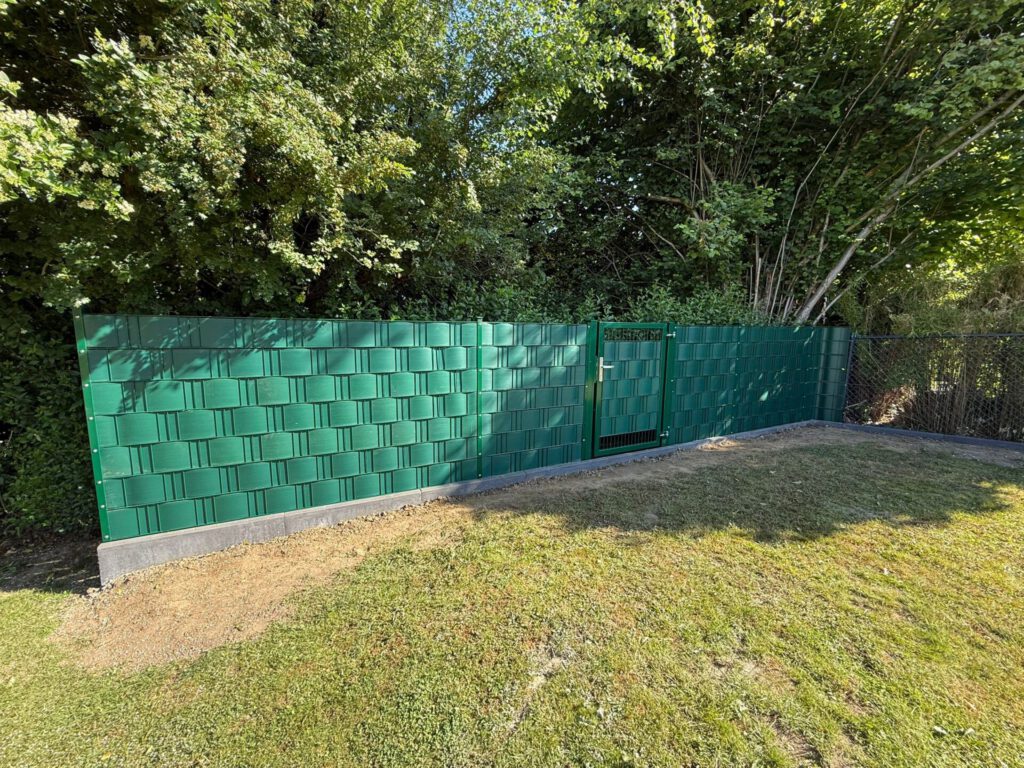 AT Garten- & Tiefbau Grüner Metallzaun mit einem Tor und einer strukturierten Oberfläche im Gartenbereich.